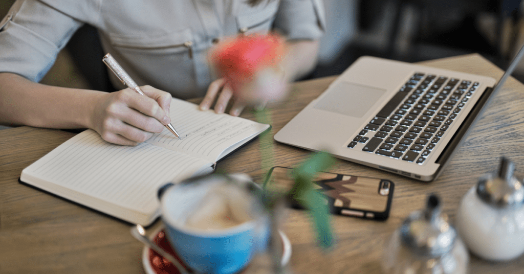 Someone writing in a notebook with laptop and phone on table working on content creation